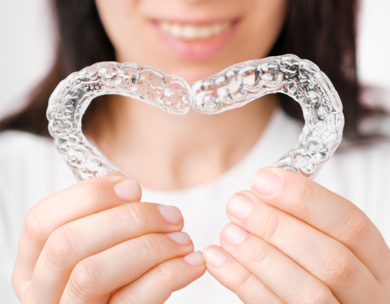 A smiling person holds two connected clear dental aligners, forming a heart shape. The background is neutral, and their expression conveys happiness.